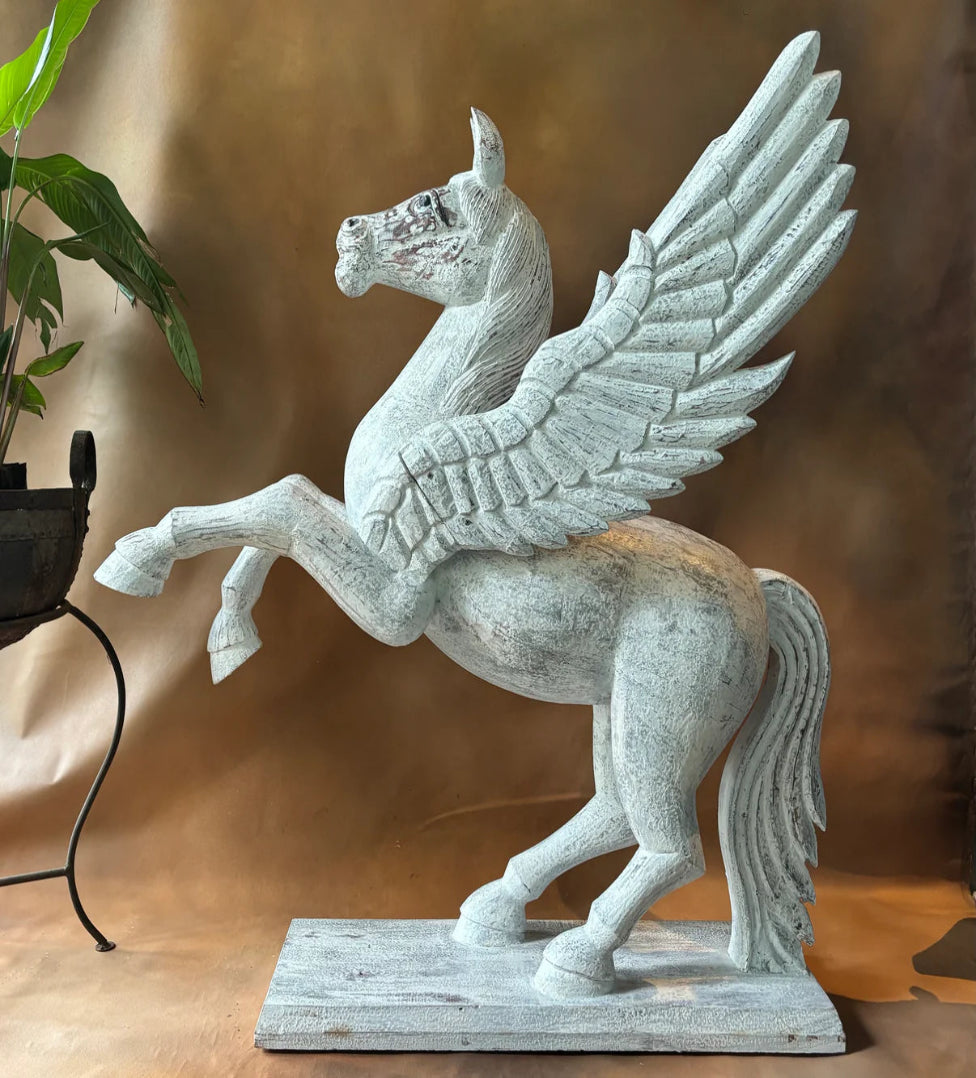A white statue of a winged horse  standing on a rectangular base with one front leg raised, displayed against a brownish background with a green plant partially visible on the left.